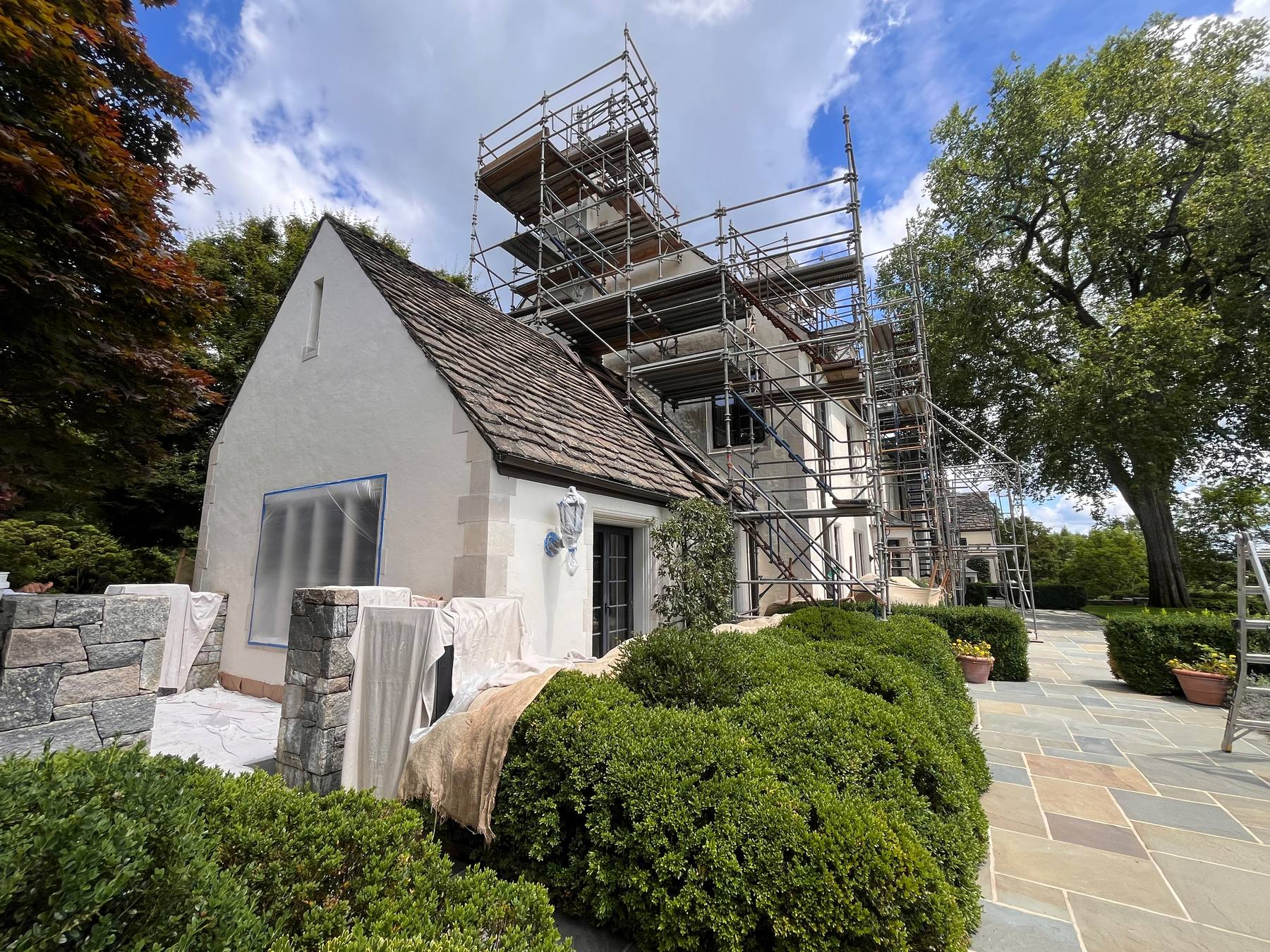 A stucco estate home with slate roof, freshly painted trim and manicured lawn, with restoration scaffolding along the chimneys.