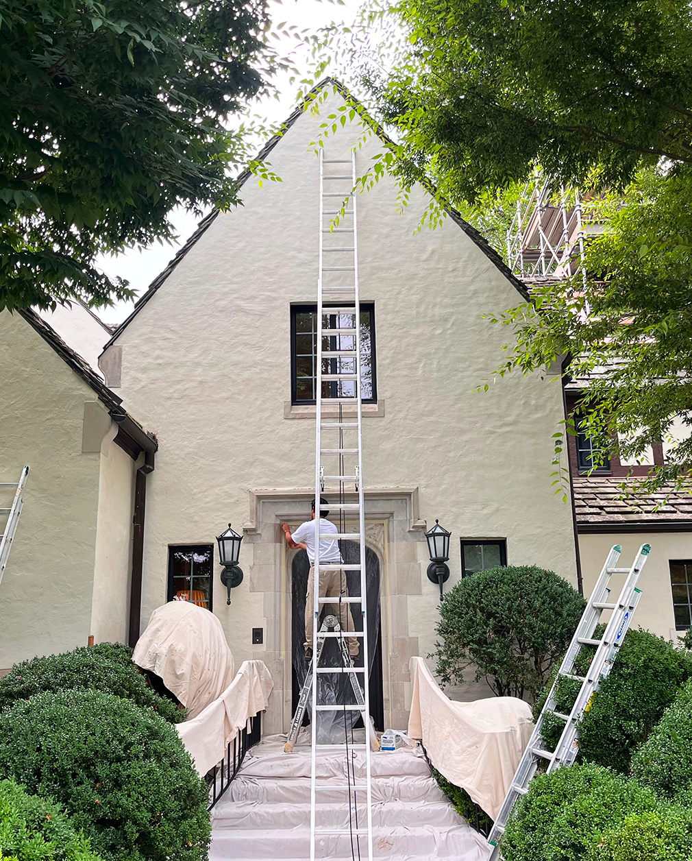 Painters on ladders restoring the stone entry of a Tudor-style estate, shrubs and railings carefully draped in canvas.